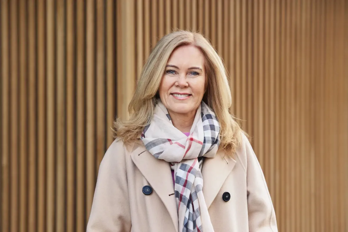 Pressebilde av kvinne i beige kåpe mot trepanel