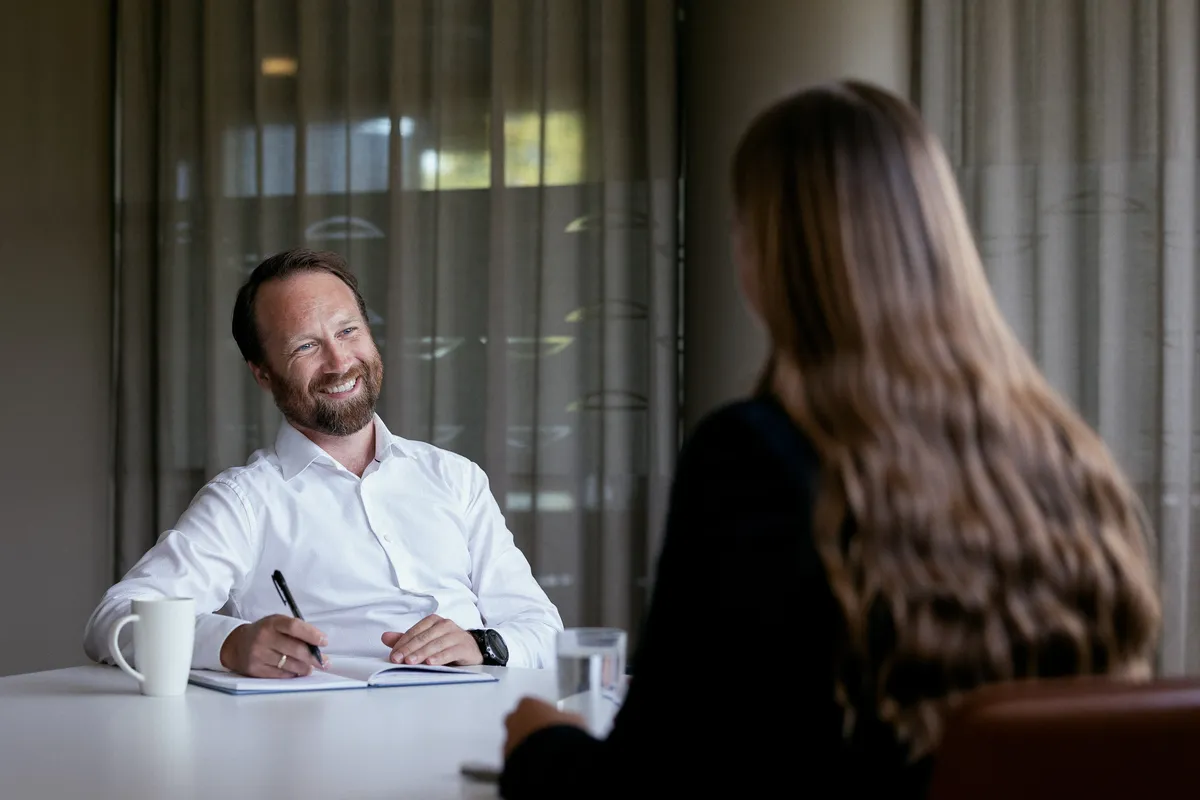 Smilende mann i hvit skjorte noterer i en blokk i et møte ved bordet, lyst kontor
